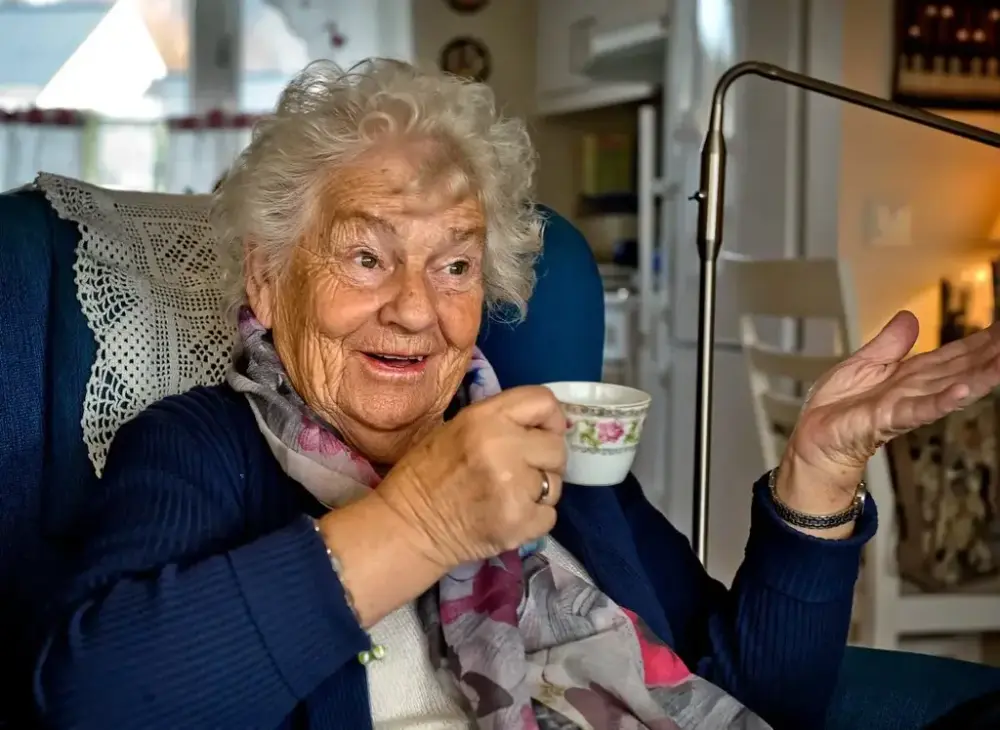 En blid eldre dame (Kirsten Tangen) sitter i en lenestol med en blomstret kaffekopp i hånda, gestikulerende. 