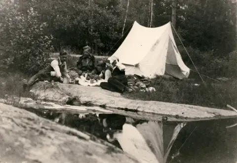 Fotografi taget från en tältplats. 
Åke Börjesson halvligger längst ut till vänster, med höger ben utsträckt. Vi ser hur de äter och hur ett gasolkök med en kastrull står ovanpå.
Okänt årtal för fotografering.