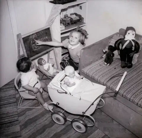 En flicka sitter på golvet och tittar upp mot fotografens håll, när bilden tas. En dockvagn med en docka i, står parkerad i närheten av spädbarnet som sitter i barnstolen på golvet. 
En soffa står till höger i bild och en docka föreställande en tomte med röd luva ligger i ett hörn. Ett dockskåp står bredvid flickan och på golvet ligger två stycken mattor, snett överlappande varandra.