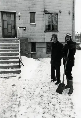 Ett eternithus i två våningar syns i vinterlandskapet. Det går tydligt att se husnumret fem uppsatt bredvid entrédörren. Männen som står bredvid varandra är Christina Lernbrinks make och en granne till paret Lernbrink. 

Osäkert när fotografiet är taget.