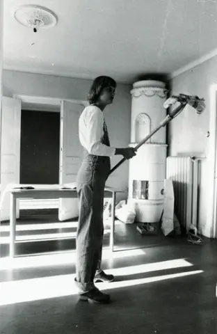 Christina Lernbrink städar inomhus. Ett stort gammaldags värmeelement står placerat bredvid kakelugnen, till höger i bild. 

Osäkert när fotografiet är taget.