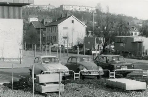 Bosgårdsgatan 5 på avstånd. I förgrunden ser vi tre personbilar parkerade: En Saab, en Volkswagen samt en Volvo. På den lilla vita skylten står texten: vårdcentral huvudentré. En arbetarbarack finns utplacerad till höger i bild. Vi ser också kantstenar som används för trottoarkanter, ligga samlade. 
På Störtfjället i bakgrunden ser vi delar av området Enerbacken.

Osäkert när fotografiet är taget.
