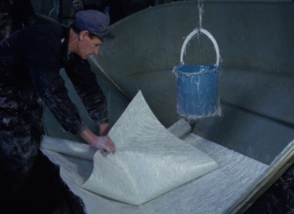 Färgfilm ur Monarks arkiv som visar företagets tillverkning av fritidsbåtar i glasfiberarmerad plast.


En träkonstruktion spänns fast med tvingar. En uppochnedvänd skrovform filmas. 


Två sorters glasfiberdukar visas för kameran: en slätare glasfiberväv och en grövre glasfibermatta. På ett långt bord dras längder av glasfibermatta ut från en stor rulle. De kapas i stycken som lämnas på hög. En form markeras på den översta mattan med hjälp av en kartongmall, och när högen lagts till rätta används en såg för att kapa formen genom alla mattor samtidigt.


Insidan av en tredimensionell skrovmall putsas och sprejas blå. En formskuren glasfibermatta läggs i mallen, rullas ut och justeras så att den täcker halva skrovet. Två arbetare använder målarrollers för att dränka in glasfibrerna i flytande plast – troligtvis polyester. En mindre metallroller används senare för att säkerställa att duken sluter tätt i mallens alla hörn och kanter.


I resten av verkstaden tillverkas fler båtdelar med samma metoder. I exempelvis toft och skott plastas även träplankor och marinplyfa in i konstruktionen som distansmaterial. En maskin används för att spreja glasfiber på en konvex yta som sedan täcks av ett lager polyester. 


I ett skrov fästs en tillskuren bit marinplyfa med klamrar mot insidan av akterspegeln. Dess form agerar mall för urtag som görs med sticksåg i akterns överkant, varefter den råa trä- och fiberkanten slipas med slipmaskin. Några skumplattor läggs ovanpå en båts aktertoft. Ett skrov lyfts upp ur sin form så att dess blåa exteriör blir synlig. Insidan av skrovet sprejas med grå färg och ytbehandlas.


Skrovet rullas iväg och vi går vidare till tillverkning av trädetaljer. Plankor spänns fast i böjda formar, flera tofter hänger på rad för att limmet i fogarna ska torka och en durk spikas ihop. Träet ytbehandlas och läggs att torka innan det fästs på de glasfiberskroven med borr och skruv. Färdigställda och inplastade båstskrov står uppradade utomhus på en snöig plan.


När ett hål i skrovet ska repareras används först en sticksåg för att kapa bort de fransade kanterna av glasfiberväven. Kanterna på det nya hålet putsas med en slipmaskin. En metallbit tejpas fast över hålet från skrovets utsida, så att det från insidan går att applicera ett nytt lager polyester med pensel. Ny glasfibermatta läggs på över plasten och täcks med ytterligare polyester. När lagningen härdat, metallen plockas bort och båtsidan ytbehandlats är lagningen så gott som osynlig.


Slutligen visas två färdigställda fritidsbåtar som hoppar fram i hög hastighet över vågigt vatten.