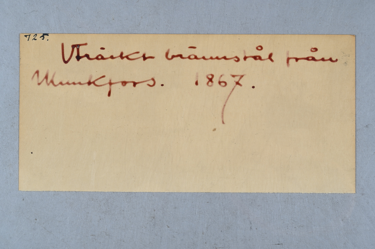 Prov på uträckt brännstål från Munkfors bruk. Två tillhörande lappar med notering: "725. Uträckt brännstål från Munkfors. 1867." Märkning på provet med notering: "AEB."