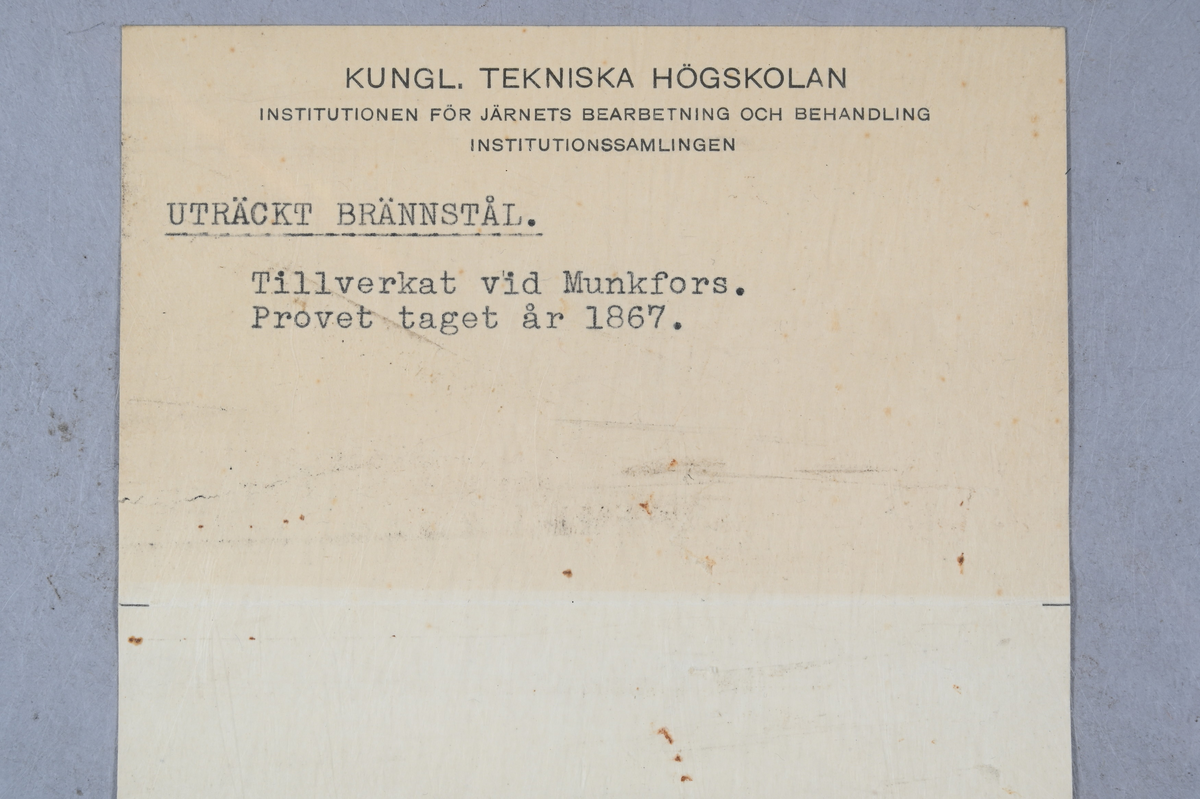 Prov på uträckt brännstål från Munkfors bruk. Två tillhörande lappar med notering: "725. Uträckt brännstål från Munkfors. 1867." Märkning på provet med notering: "AEB."