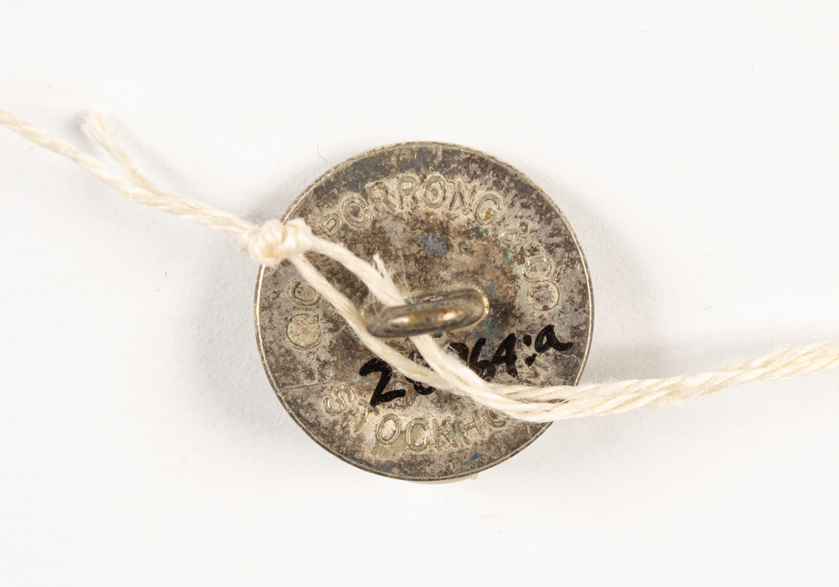 2 st cirkelrunda knappar i silver med SAL:s monogram och utmed kanten flätmönter, gjutet.

På baksidan pålödd ring för fäste på plagg, text: C.C. SPORRONG & CO STOCKHOLM