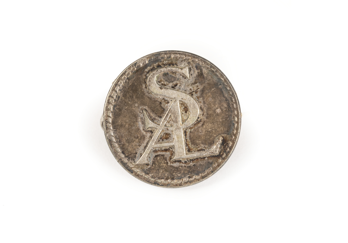 2 st cirkelrunda knappar i silver med SAL:s monogram och utmed kanten flätmönter, gjutet.

På baksidan pålödd ring för fäste på plagg, text: C.C. SPORRONG & CO STOCKHOLM