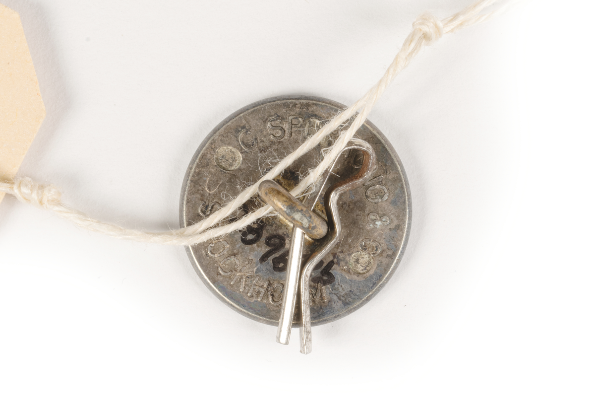 2 st cirkelrunda knappar i silver med SAL:s monogram och utmed kanten flätmönter, gjutet.

På baksidan pålödd ring för fäste på plagg, text: C.C. SPORRONG & CO STOCKHOLM