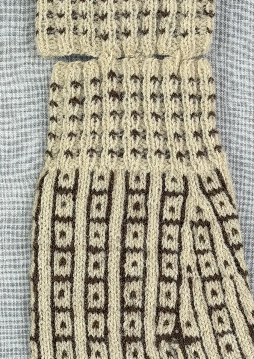 En stickad fingervante, med mönstrad mudd i 2-2 resår (2 räta och 2 aviga maskor) och mönsterstickad vante i slätstickning, av 2-trådigt ullgarn.