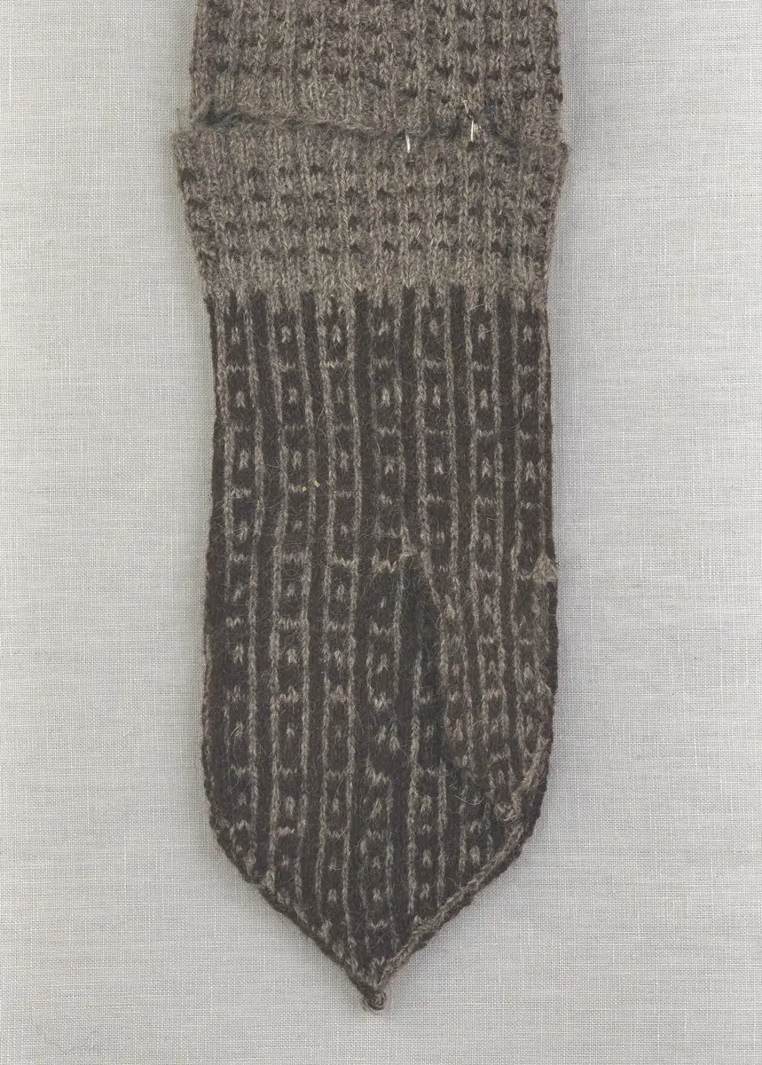 En stickad fingervante, med mönstrad mudd i 2-2 resår (2 räta och 2 aviga maskor) och mönsterstickad vante i slätstickning, av 2-trådigt ullgarn.