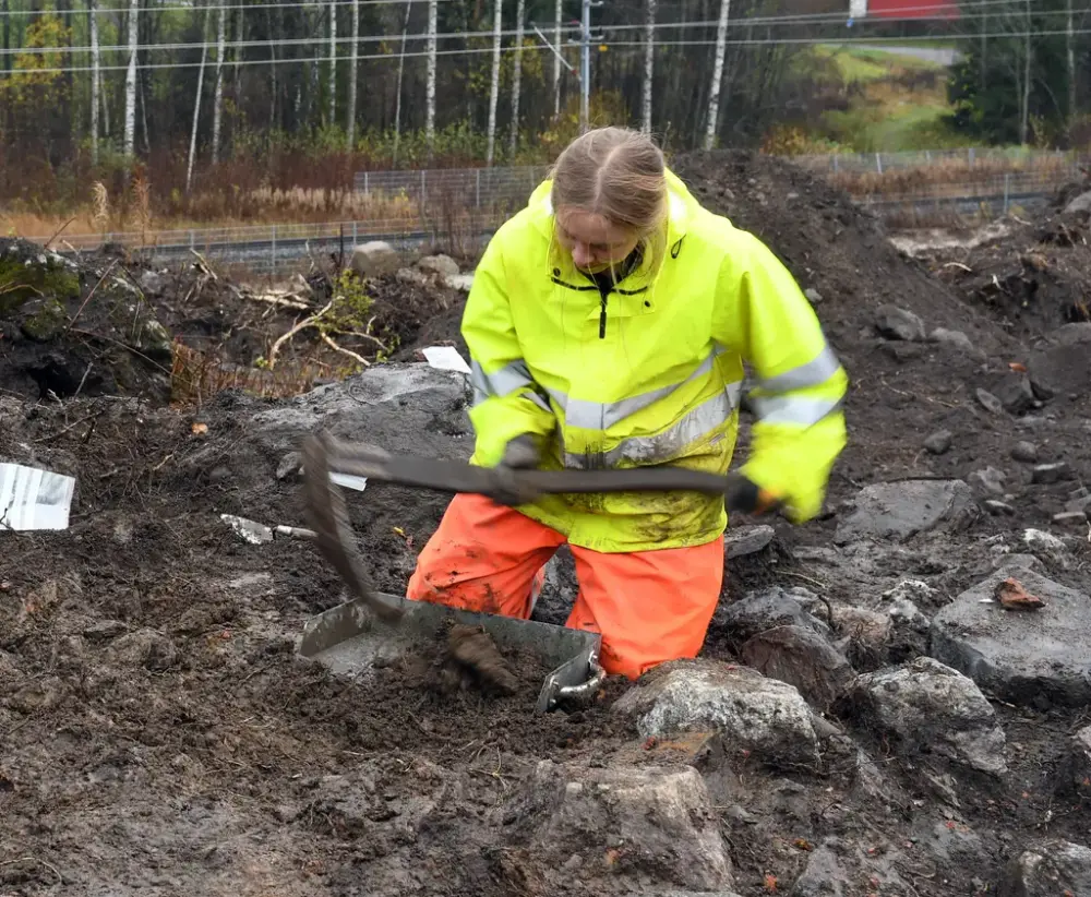 En kvinna i gul reflexväst och orangea byxor sitter på knä i jorden med ett arbetsredskap i handen och en skyffel på backen.