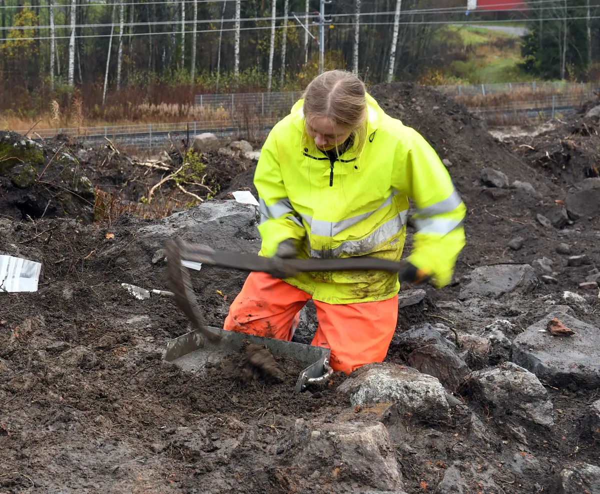 En kvinna i gul reflexväst och orangea byxor sitter på knä i jorden med ett arbetsredskap i handen och en skyffel på backen.