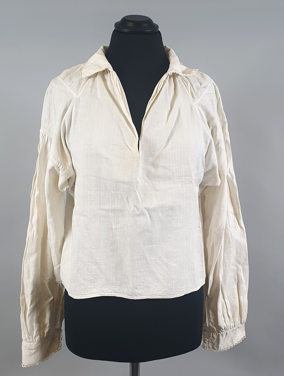 Bunadsskjorte av lin, med krage og mansjetter med nupereller. Rysjer langs halskanten, og innlegg på skuldrene.