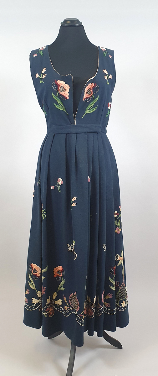 Mørkeblå bunad av ull med samme stoff i liv og stakk. Stakk og liv er sydd sammen med smal linning i livet. Foldene er lagt i samme retning langs sømmen til linningen. Broderte blomster med blader i grupper både på forstykkene, ryggen og spredt utover stakken. Hvit, bølget bord med franske knuter langs skjørtekanten. Brodert med ulltråd og metalltråd. Trykknapper i åpning foran på stakken og metallhemper foran på livet.