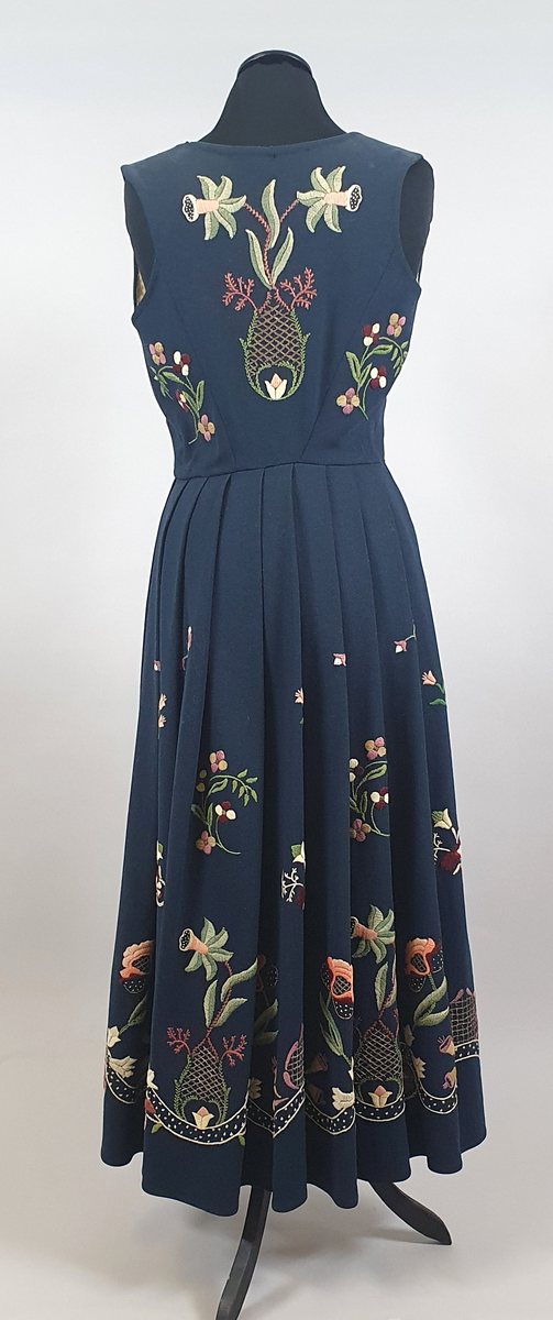 Mørkeblå bunad av ull med samme stoff i liv og stakk. Stakk og liv er sydd sammen med smal linning i livet. Foldene er lagt i samme retning langs sømmen til linningen. Broderte blomster med blader i grupper både på forstykkene, ryggen og spredt utover stakken. Hvit, bølget bord med franske knuter langs skjørtekanten. Brodert med ulltråd og metalltråd. Trykknapper i åpning foran på stakken og metallhemper foran på livet.