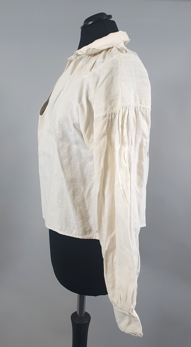 Bunadsskjorte av lin, med krage og mansjetter med nupereller. Rysjer langs halskanten, og innlegg på skuldrene.