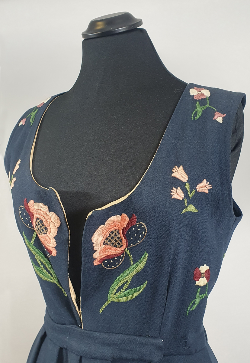 Mørkeblå bunad av ull med samme stoff i liv og stakk. Stakk og liv er sydd sammen med smal linning i livet. Foldene er lagt i samme retning langs sømmen til linningen. Broderte blomster med blader i grupper både på forstykkene, ryggen og spredt utover stakken. Hvit, bølget bord med franske knuter langs skjørtekanten. Brodert med ulltråd og metalltråd. Trykknapper i åpning foran på stakken og metallhemper foran på livet.