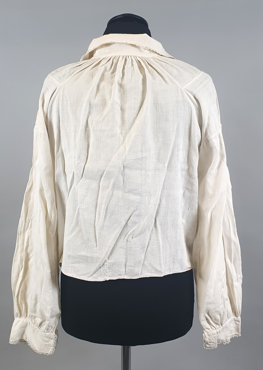 Bunadsskjorte av lin, med krage og mansjetter med nupereller. Rysjer langs halskanten, og innlegg på skuldrene.