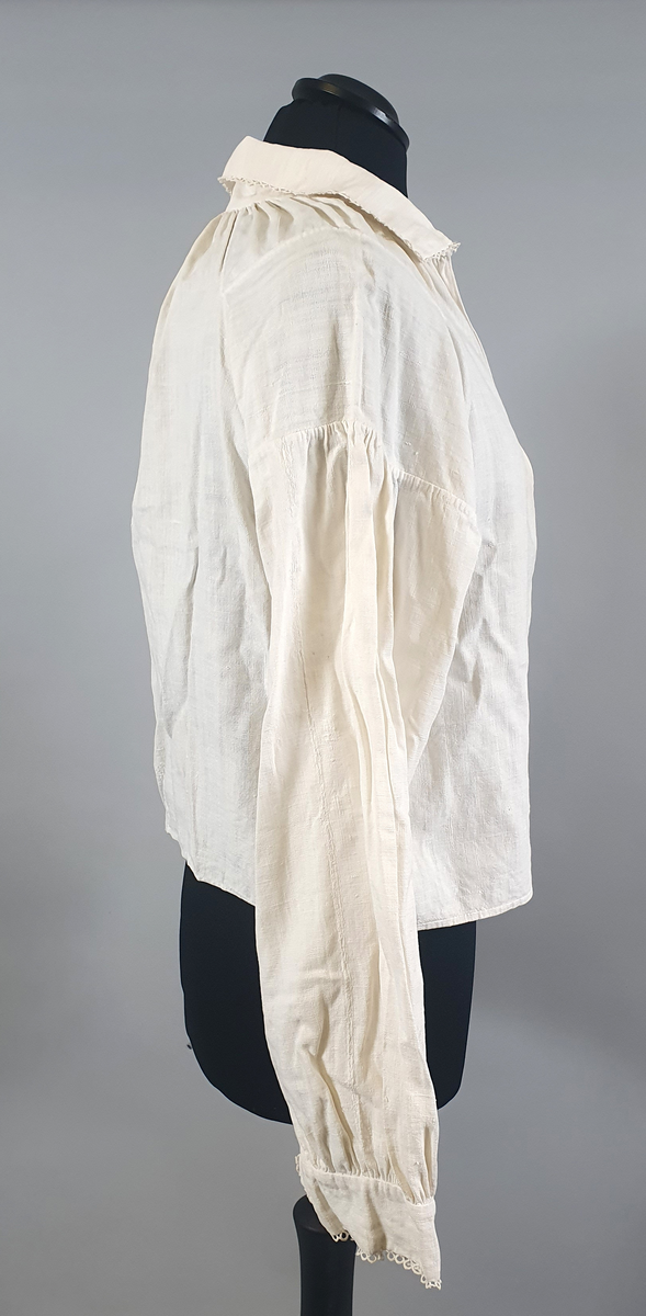Bunadsskjorte av lin, med krage og mansjetter med nupereller. Rysjer langs halskanten, og innlegg på skuldrene.