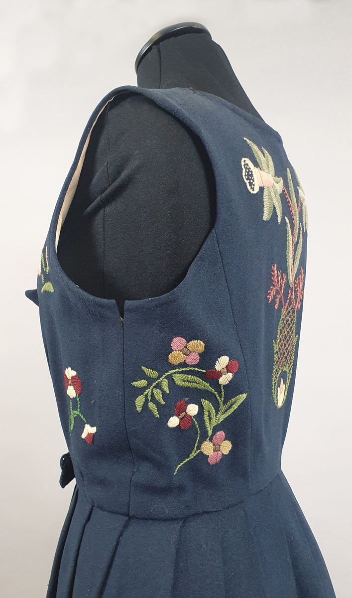 Mørkeblå bunad av ull med samme stoff i liv og stakk. Stakk og liv er sydd sammen med smal linning i livet. Foldene er lagt i samme retning langs sømmen til linningen. Broderte blomster med blader i grupper både på forstykkene, ryggen og spredt utover stakken. Hvit, bølget bord med franske knuter langs skjørtekanten. Brodert med ulltråd og metalltråd. Trykknapper i åpning foran på stakken og metallhemper foran på livet.