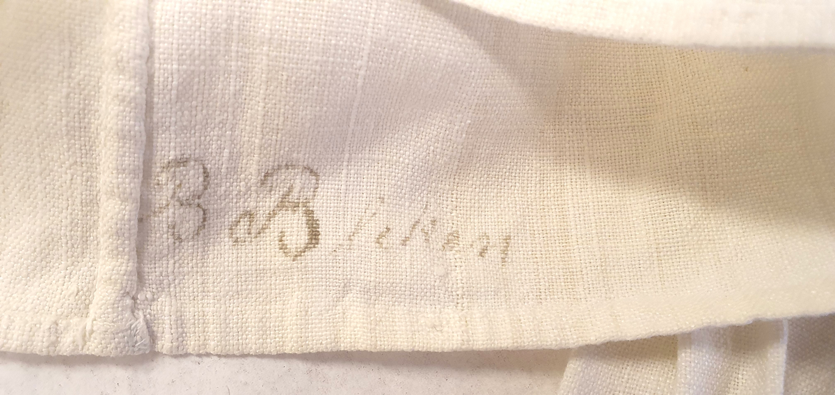 Bunadsskjorte av lin, med krage og mansjetter med nupereller. Rysjer langs halskanten, og innlegg på skuldrene.