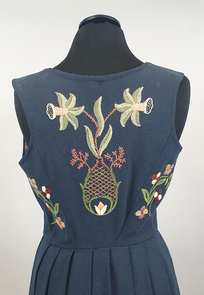 Mørkeblå bunad av ull med samme stoff i liv og stakk. Stakk og liv er sydd sammen med smal linning i livet. Foldene er lagt i samme retning langs sømmen til linningen. Broderte blomster med blader i grupper både på forstykkene, ryggen og spredt utover stakken. Hvit, bølget bord med franske knuter langs skjørtekanten. Brodert med ulltråd og metalltråd. Trykknapper i åpning foran på stakken og metallhemper foran på livet.