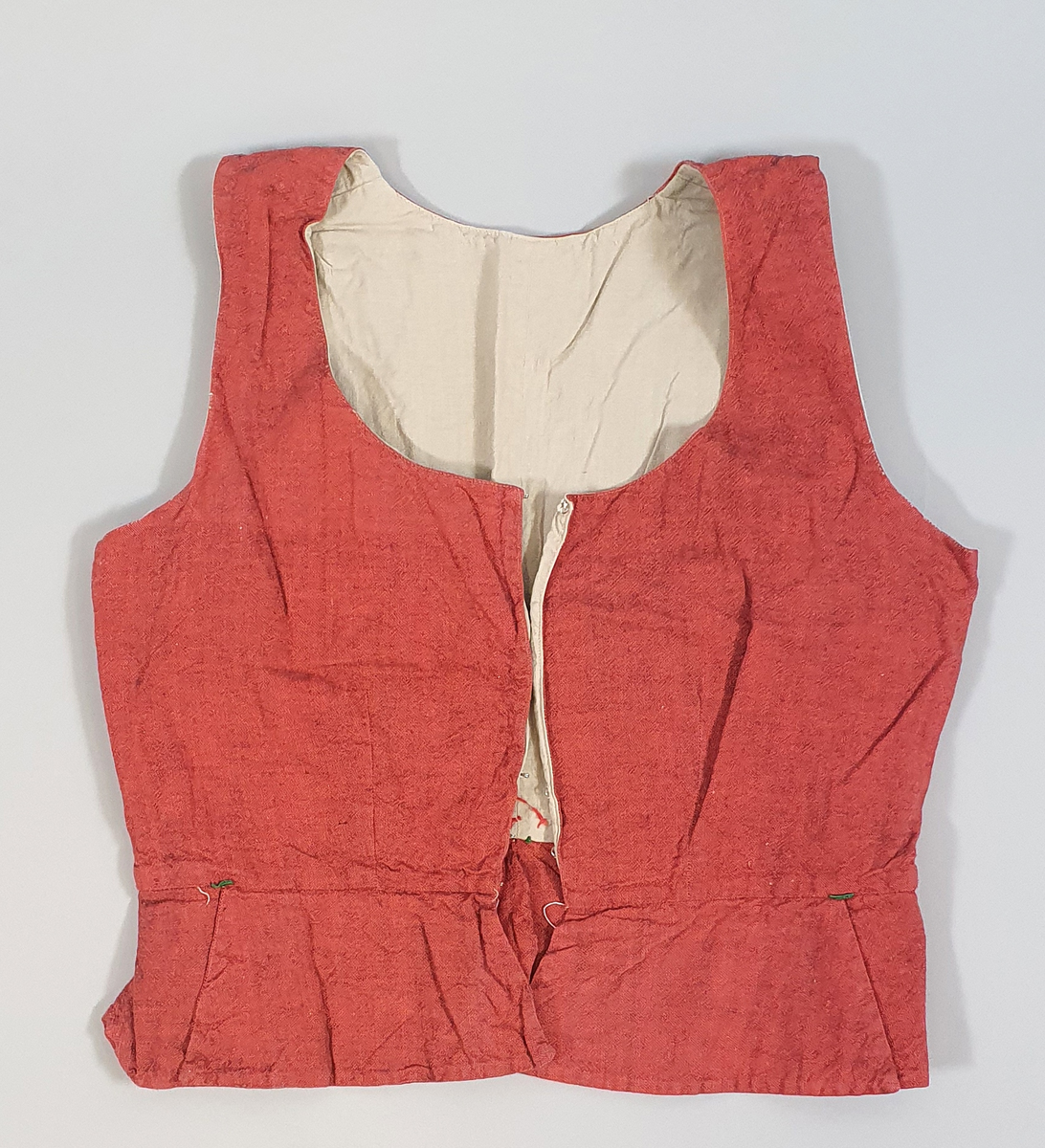 Rødt liv av lin, med seks skjøter (flak) somskår ut fra livlinjen. Metallhemper i åpningen foran. Livet er fôret med ubleket bomull. Brodert navn på innsiden av forstykket.