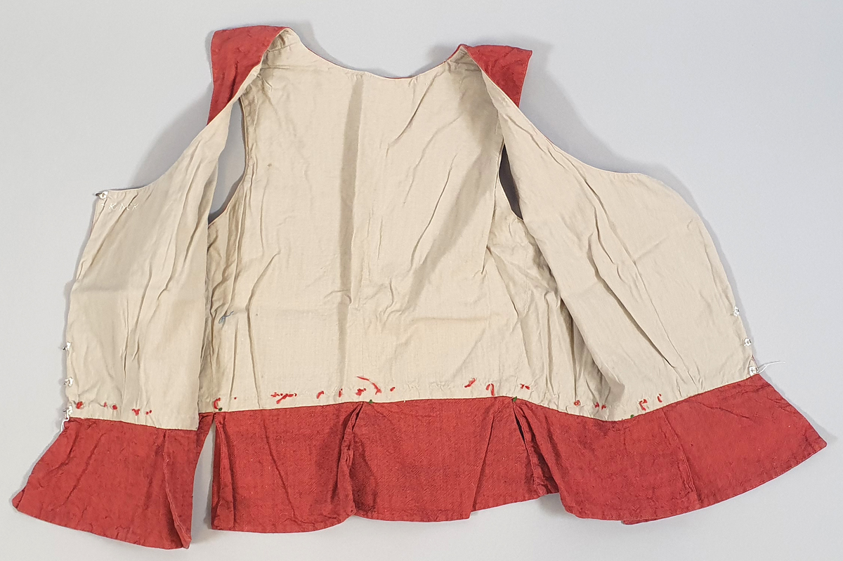 Rødt liv av lin, med seks skjøter (flak) somskår ut fra livlinjen. Metallhemper i åpningen foran. Livet er fôret med ubleket bomull. Brodert navn på innsiden av forstykket.