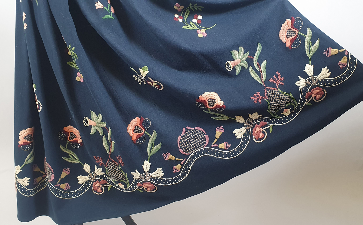 Mørkeblå bunad av ull med samme stoff i liv og stakk. Stakk og liv er sydd sammen med smal linning i livet. Foldene er lagt i samme retning langs sømmen til linningen. Broderte blomster med blader i grupper både på forstykkene, ryggen og spredt utover stakken. Hvit, bølget bord med franske knuter langs skjørtekanten. Brodert med ulltråd og metalltråd. Trykknapper i åpning foran på stakken og metallhemper foran på livet.
