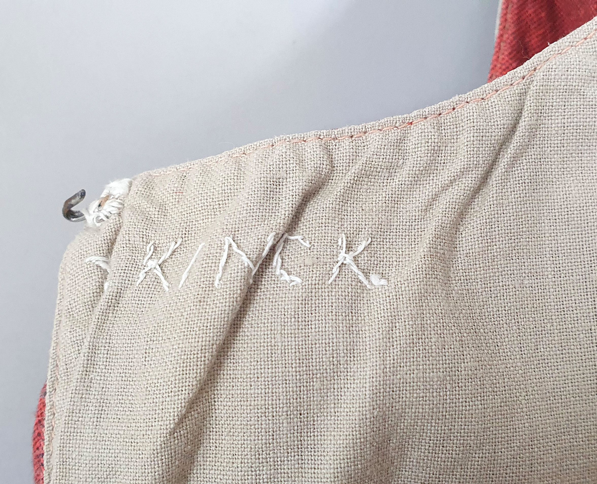 Rødt liv av lin, med seks skjøter (flak) somskår ut fra livlinjen. Metallhemper i åpningen foran. Livet er fôret med ubleket bomull. Brodert navn på innsiden av forstykket.