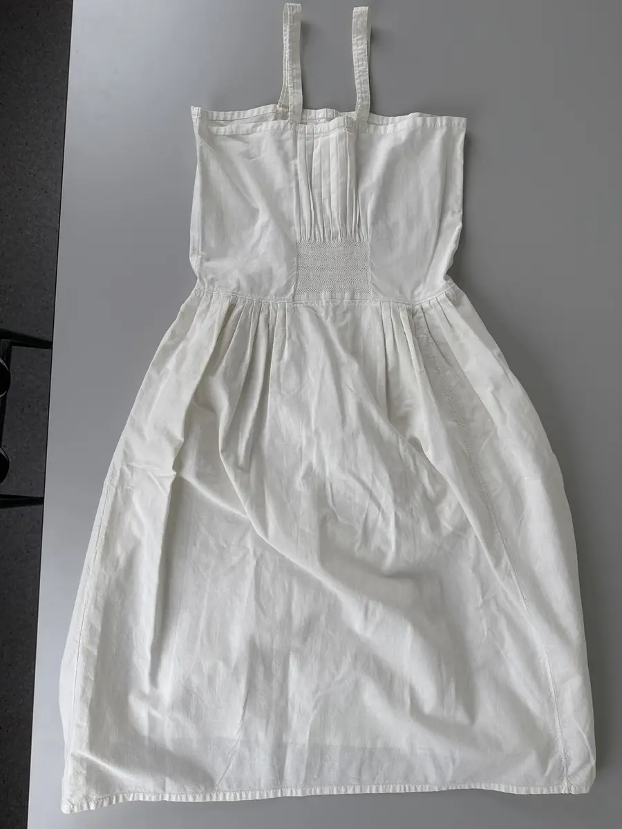 Särk, dialektalt bono eller neadel (Jonsson, Delsbodräkterna, 1977), av bomullsväv med hängslen. Övre delen av blekt bomullsväv med piprynkor och vitsöm på ett fyrkantigt fält, 8 gånger 9 cm, mitt fram och mitt bak. Lagda veck runtom utom mitt fram och mitt bak. Samma tyg i kjolen.