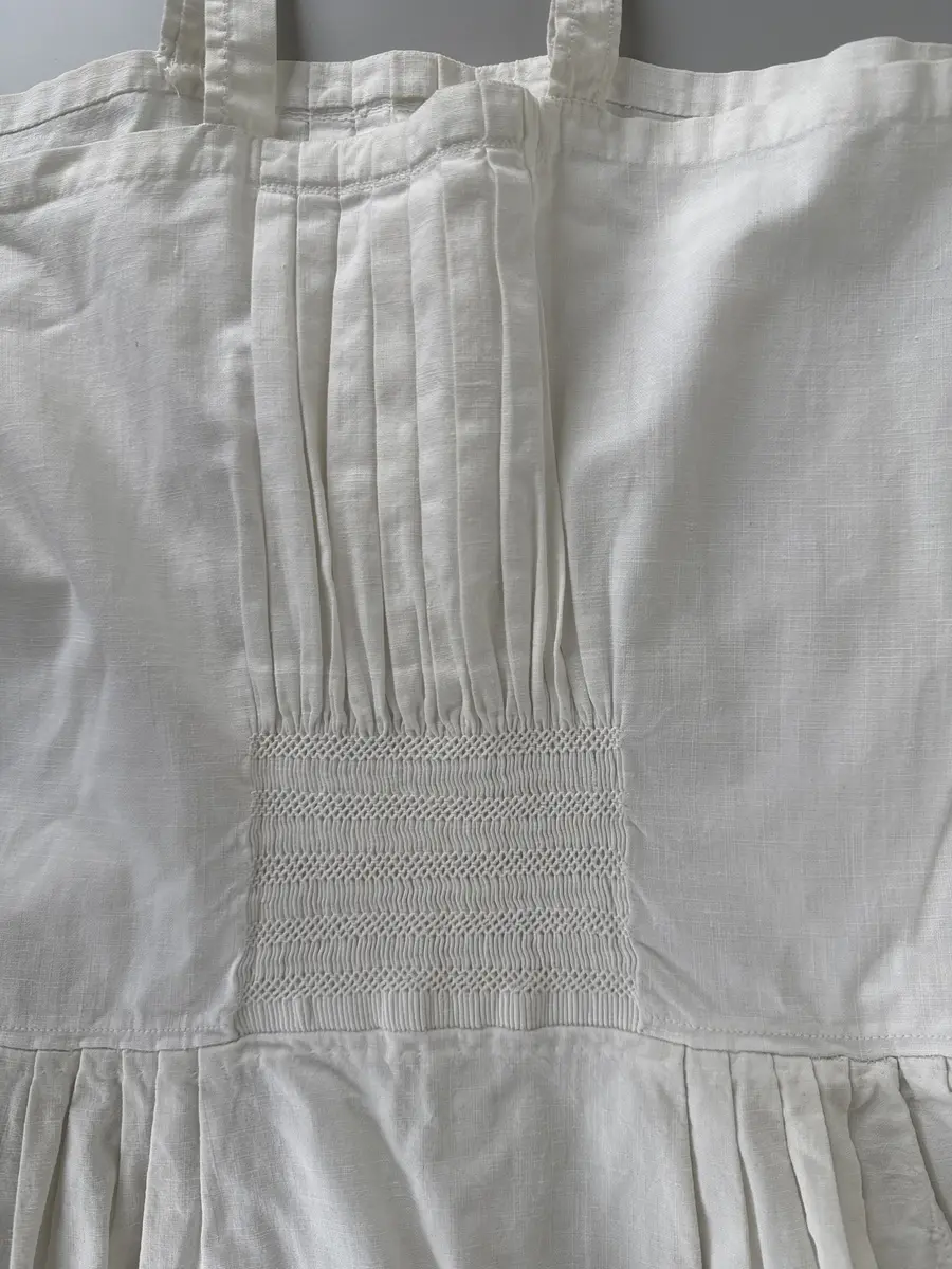 Särk, dialektalt bono eller neadel (Jonsson, Delsbodräkterna, 1977), av bomullsväv med hängslen. Övre delen av blekt bomullsväv med piprynkor och vitsöm på ett fyrkantigt fält, 8 gånger 9 cm, mitt fram och mitt bak. Lagda veck runtom utom mitt fram och mitt bak. Samma tyg i kjolen.