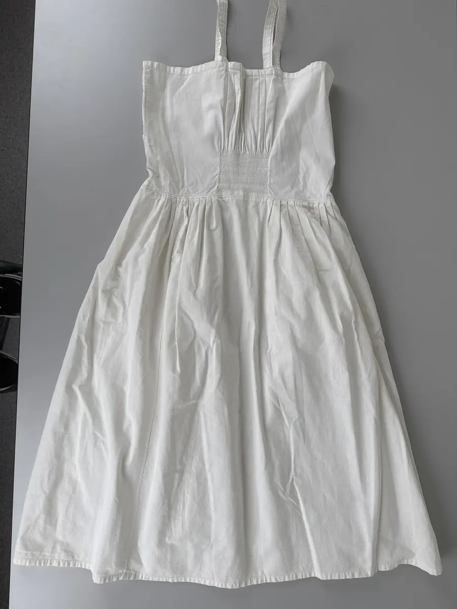 Särk, dialektalt bono eller neadel (Jonsson, Delsbodräkterna, 1977), av bomullsväv med hängslen. Övre delen av blekt bomullsväv med piprynkor och vitsöm på ett fyrkantigt fält, 8 gånger 9 cm, mitt fram och mitt bak. Lagda veck runtom utom mitt fram och mitt bak. Samma tyg i kjolen.