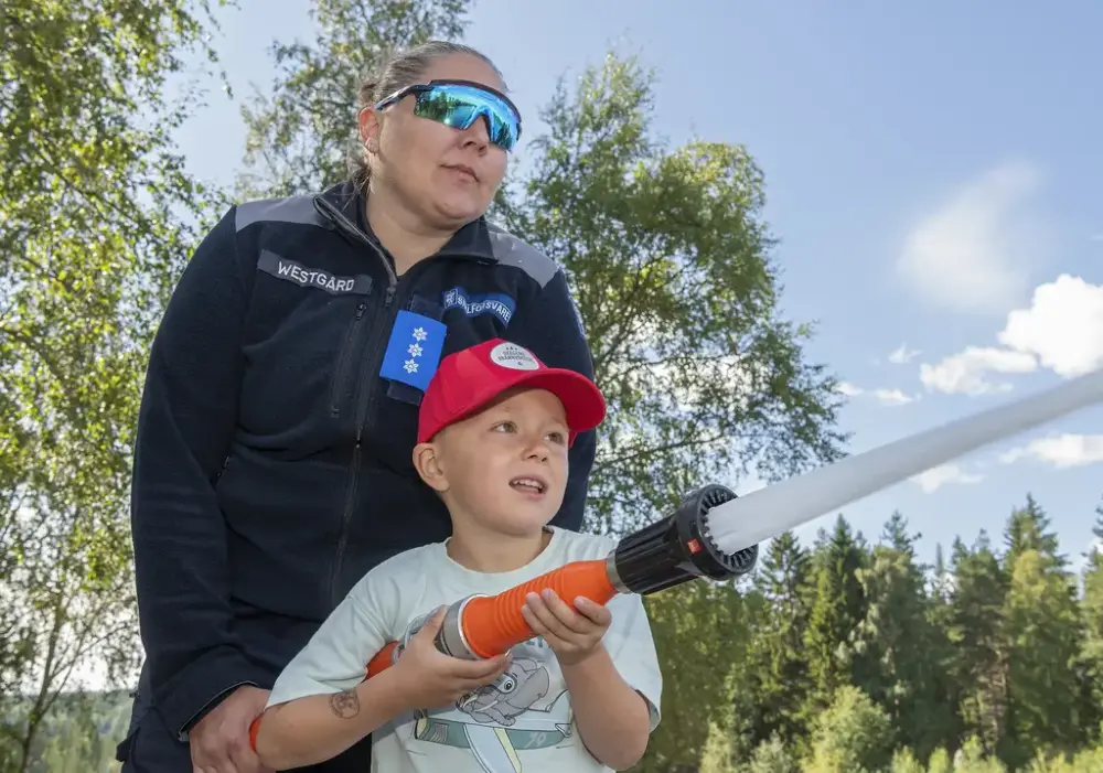 Dame fra sivilforsvaret hjelper en gutt med å spyle med en brannslange 