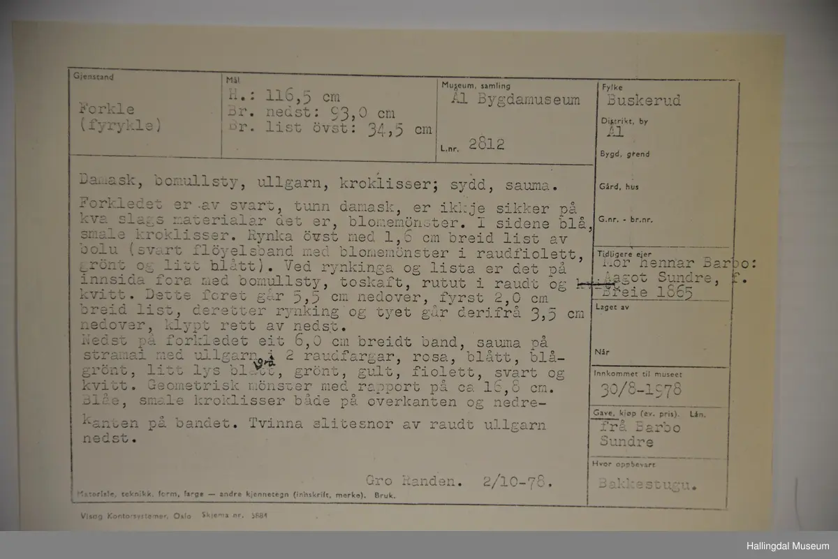 Forkledet er av svart, tunn damask, blomemønster. I sidene blå, smale kroklisser. Rynka øvst med list av bolu (svart fløyelsband med blomemønster i raudfiolett, grønt og litt blått). Ved rynkinga og lista er det på innsida fora med bomullsty, toskaft, rutut i raudt og kvitt. Nedst på forkledet eit band, sauma på stramei med ullgarn i 2 raudfargar, rosa, blått, blågrønt, litt lys blått, grønt, gult, fiolett, svart og kvitt. Geometrisk mønster. Blå, smale kroklisser både på overkanten og nedrekanten på bandet. Tvinna slitesnor av raudt ullgarn nedst.