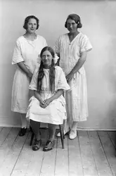 Portrett av tre kvinner. En kvinne sitter på en stol, to stå