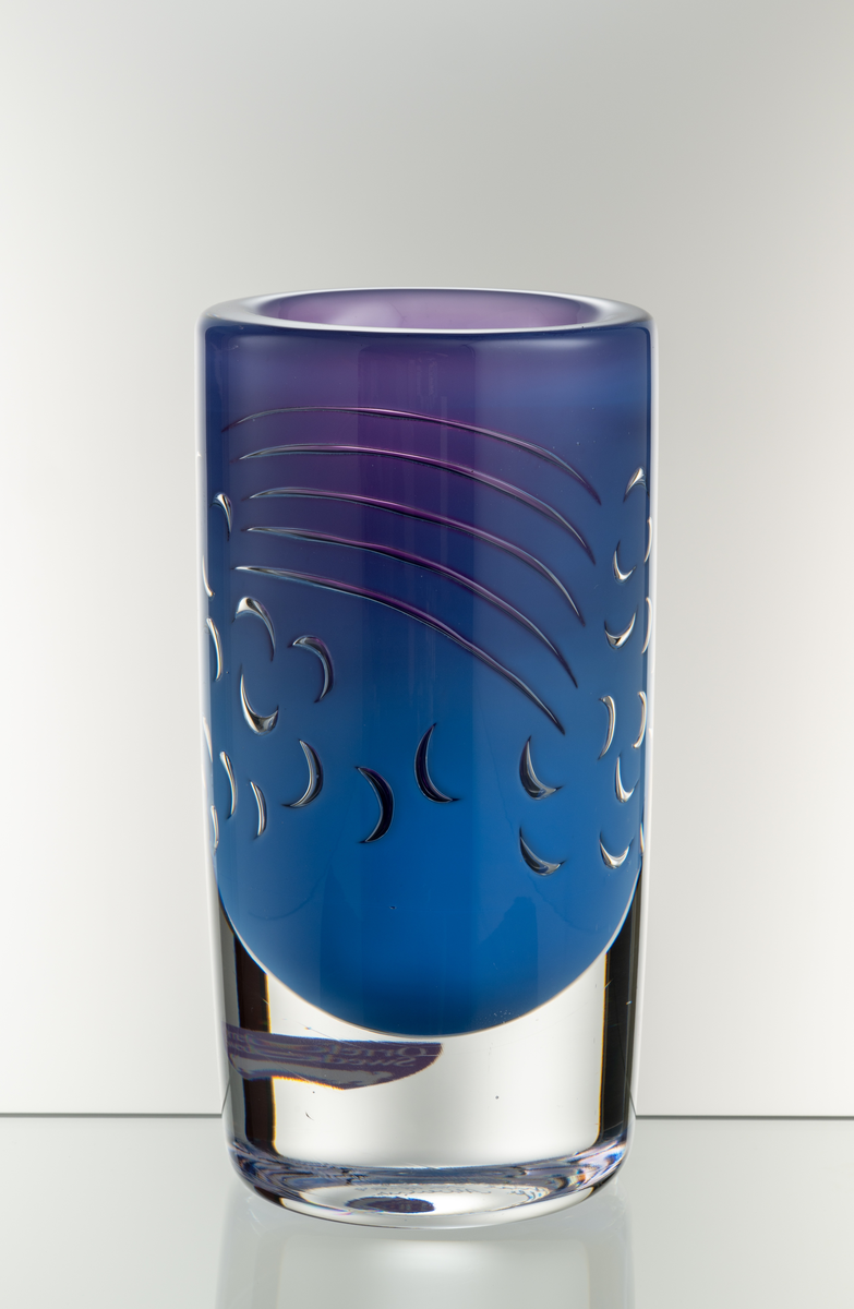 Arielvas, tjockväggig. Cylindrisk form. Ametistlila och delftblå färgton med abstrakt luftmönster i form av bågar och linjer.
Formgivare: Olle Alberius.