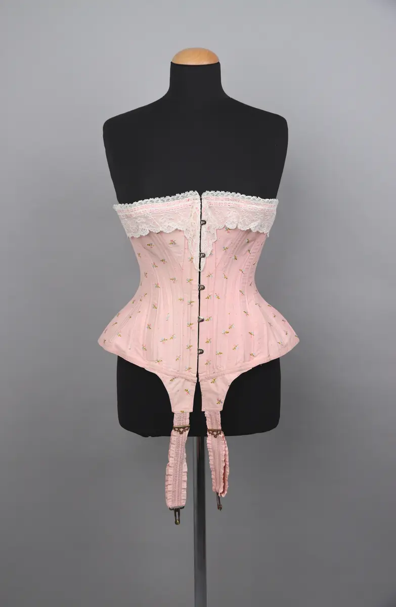 Modekorsett av ljusrosa bomull. Med ett par strumpeband i främre nederkanten. Strumpebanden har varit elastiska och har rester av torrt gummi. De är tillverkade med invävda volanger på båda sidor. Spännena på strumpebanden är i form av blomrankor och en rosett. Överkanten på korsetten är dekorerad med bred vit spets med inträdda dubbla rosa sidenband. Det rosa yttertyget har ett mönster av broderade blommor och prickar i rosa, grönt och blått. Benad runt om hela plagget. Snörning i ryggen genom 15 par snörhål med ljusrosa flätat snodd. Metallbeslag av mässing; spännen, planschett och öljetter. Insida är fodrad med stadig bomullslärft och bomullsband. Vävd etikett med tillverkarens namn i midjebandet. Stämpel på midjebandet med garanti och modell-namn. 2 pappersetiketter ev. med pris eller storleksuppgifter.