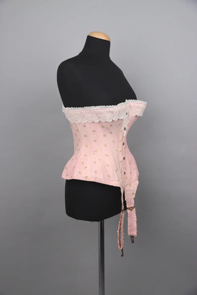 Modekorsett av ljusrosa bomull. Med ett par strumpeband i främre nederkanten. Strumpebanden har varit elastiska och har rester av torrt gummi. De är tillverkade med invävda volanger på båda sidor. Spännena på strumpebanden är i form av blomrankor och en rosett. Överkanten på korsetten är dekorerad med bred vit spets med inträdda dubbla rosa sidenband. Det rosa yttertyget har ett mönster av broderade blommor och prickar i rosa, grönt och blått. Benad runt om hela plagget. Snörning i ryggen genom 15 par snörhål med ljusrosa flätat snodd. Metallbeslag av mässing; spännen, planschett och öljetter. Insida är fodrad med stadig bomullslärft och bomullsband. Vävd etikett med tillverkarens namn i midjebandet. Stämpel på midjebandet med garanti och modell-namn. 2 pappersetiketter ev. med pris eller storleksuppgifter.