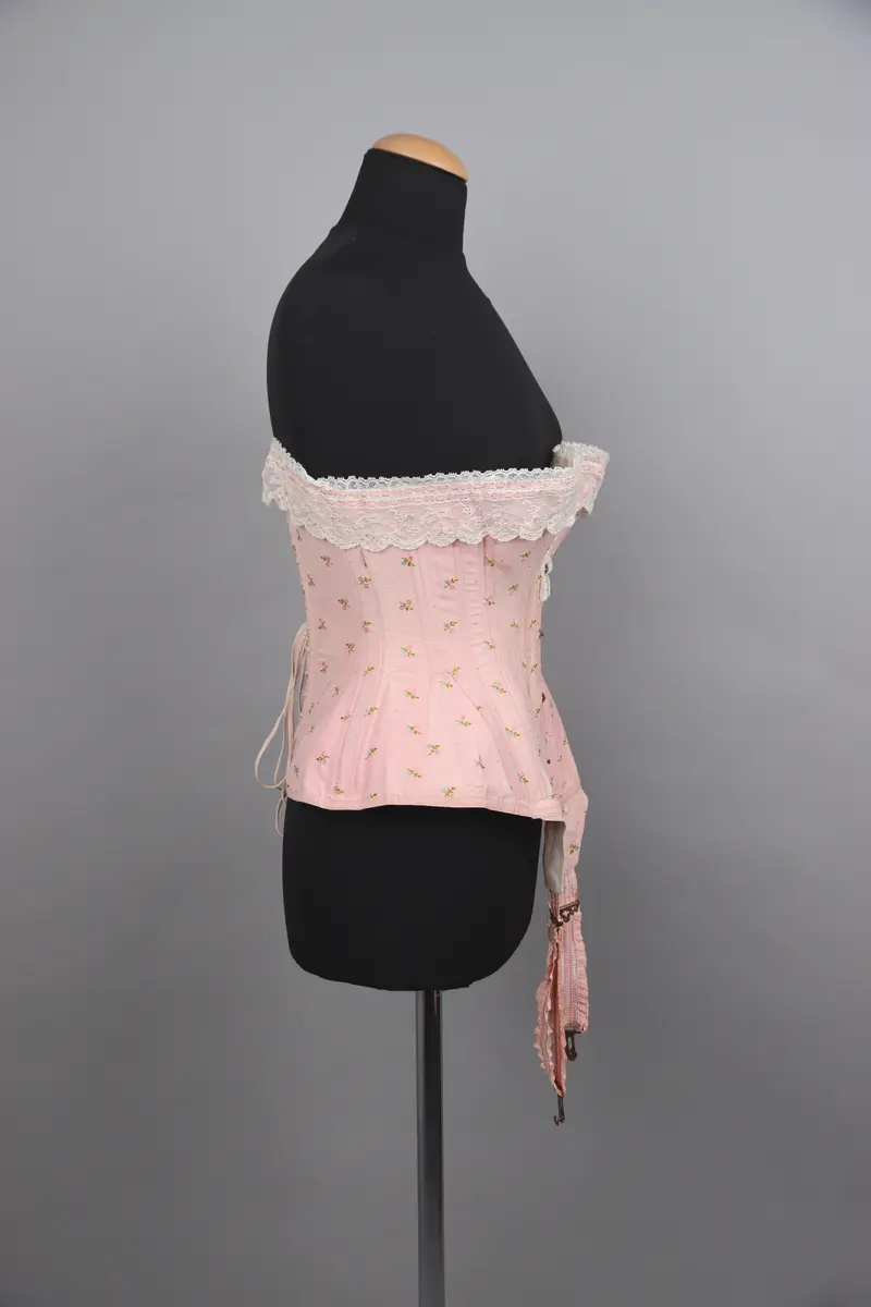 Modekorsett av ljusrosa bomull. Med ett par strumpeband i främre nederkanten. Strumpebanden har varit elastiska och har rester av torrt gummi. De är tillverkade med invävda volanger på båda sidor. Spännena på strumpebanden är i form av blomrankor och en rosett. Överkanten på korsetten är dekorerad med bred vit spets med inträdda dubbla rosa sidenband. Det rosa yttertyget har ett mönster av broderade blommor och prickar i rosa, grönt och blått. Benad runt om hela plagget. Snörning i ryggen genom 15 par snörhål med ljusrosa flätat snodd. Metallbeslag av mässing; spännen, planschett och öljetter. Insida är fodrad med stadig bomullslärft och bomullsband. Vävd etikett med tillverkarens namn i midjebandet. Stämpel på midjebandet med garanti och modell-namn. 2 pappersetiketter ev. med pris eller storleksuppgifter.