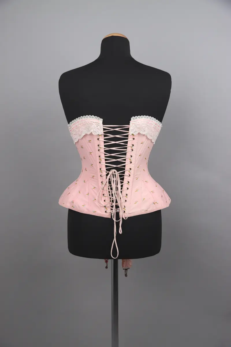 Modekorsett av ljusrosa bomull. Med ett par strumpeband i främre nederkanten. Strumpebanden har varit elastiska och har rester av torrt gummi. De är tillverkade med invävda volanger på båda sidor. Spännena på strumpebanden är i form av blomrankor och en rosett. Överkanten på korsetten är dekorerad med bred vit spets med inträdda dubbla rosa sidenband. Det rosa yttertyget har ett mönster av broderade blommor och prickar i rosa, grönt och blått. Benad runt om hela plagget. Snörning i ryggen genom 15 par snörhål med ljusrosa flätat snodd. Metallbeslag av mässing; spännen, planschett och öljetter. Insida är fodrad med stadig bomullslärft och bomullsband. Vävd etikett med tillverkarens namn i midjebandet. Stämpel på midjebandet med garanti och modell-namn. 2 pappersetiketter ev. med pris eller storleksuppgifter.