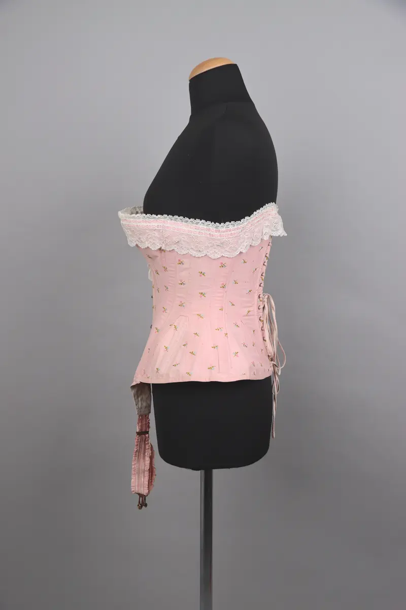 Modekorsett av ljusrosa bomull. Med ett par strumpeband i främre nederkanten. Strumpebanden har varit elastiska och har rester av torrt gummi. De är tillverkade med invävda volanger på båda sidor. Spännena på strumpebanden är i form av blomrankor och en rosett. Överkanten på korsetten är dekorerad med bred vit spets med inträdda dubbla rosa sidenband. Det rosa yttertyget har ett mönster av broderade blommor och prickar i rosa, grönt och blått. Benad runt om hela plagget. Snörning i ryggen genom 15 par snörhål med ljusrosa flätat snodd. Metallbeslag av mässing; spännen, planschett och öljetter. Insida är fodrad med stadig bomullslärft och bomullsband. Vävd etikett med tillverkarens namn i midjebandet. Stämpel på midjebandet med garanti och modell-namn. 2 pappersetiketter ev. med pris eller storleksuppgifter.