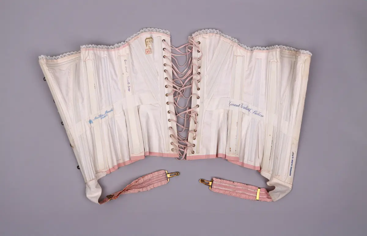 Modekorsett av ljusrosa bomull. Med ett par strumpeband i främre nederkanten. Strumpebanden har varit elastiska och har rester av torrt gummi. De är tillverkade med invävda volanger på båda sidor. Spännena på strumpebanden är i form av blomrankor och en rosett. Överkanten på korsetten är dekorerad med bred vit spets med inträdda dubbla rosa sidenband. Det rosa yttertyget har ett mönster av broderade blommor och prickar i rosa, grönt och blått. Benad runt om hela plagget. Snörning i ryggen genom 15 par snörhål med ljusrosa flätat snodd. Metallbeslag av mässing; spännen, planschett och öljetter. Insida är fodrad med stadig bomullslärft och bomullsband. Vävd etikett med tillverkarens namn i midjebandet. Stämpel på midjebandet med garanti och modell-namn. 2 pappersetiketter ev. med pris eller storleksuppgifter.