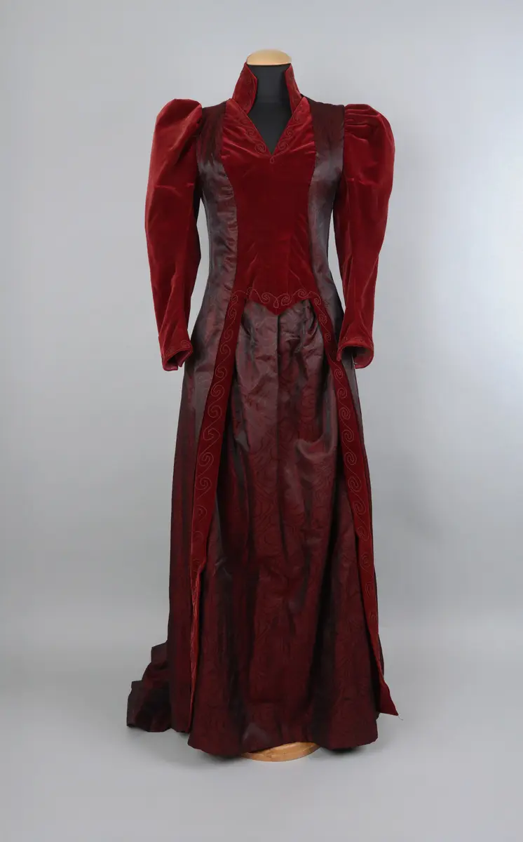 Klänning, av modell från 1891. Festklänning för dam av vinröd sammet och moire-liknande tyg. Sitter ihop i ett stycke med kjol och robe/överdel sammansatta. Stängs i ryggen med hyskor och hakar. Den höga kragen knäpps på med tryckknappar. Klänningens kjol har ett kortare släp.

