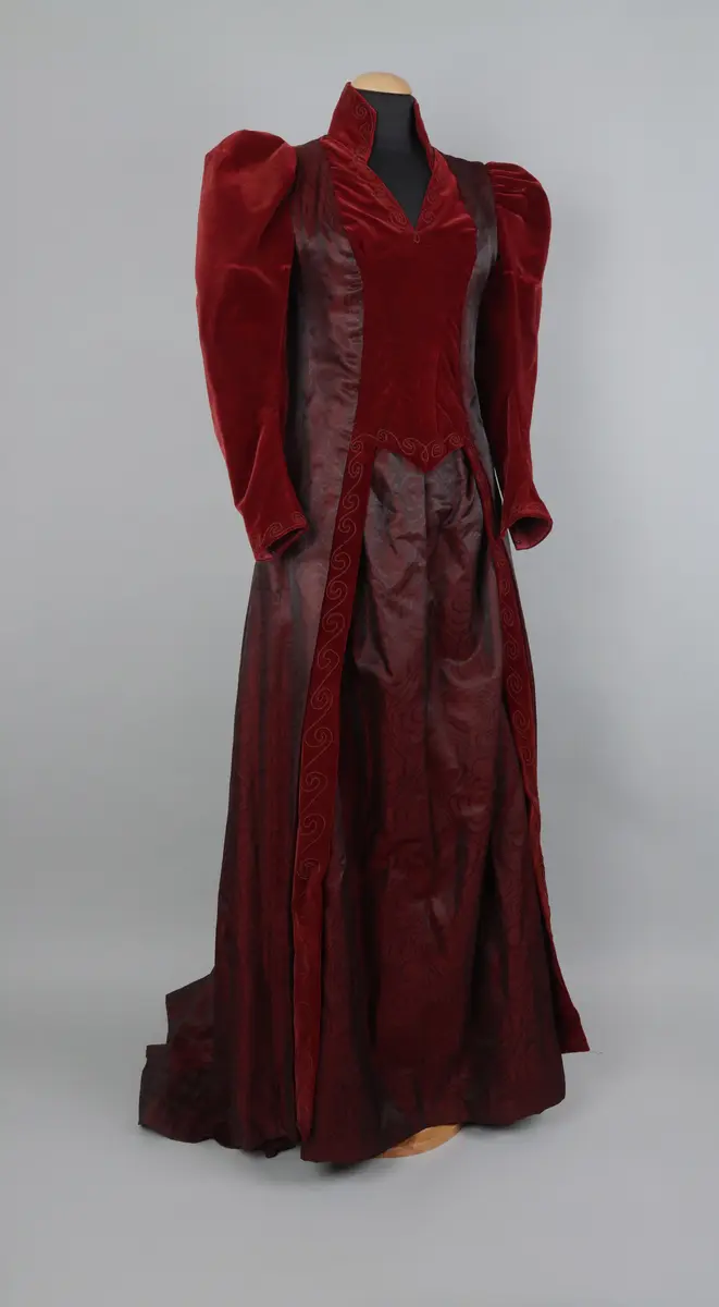 Klänning, av modell från 1891. Festklänning för dam av vinröd sammet och moire-liknande tyg. Sitter ihop i ett stycke med kjol och robe/överdel sammansatta. Stängs i ryggen med hyskor och hakar. Den höga kragen knäpps på med tryckknappar. Klänningens kjol har ett kortare släp.

