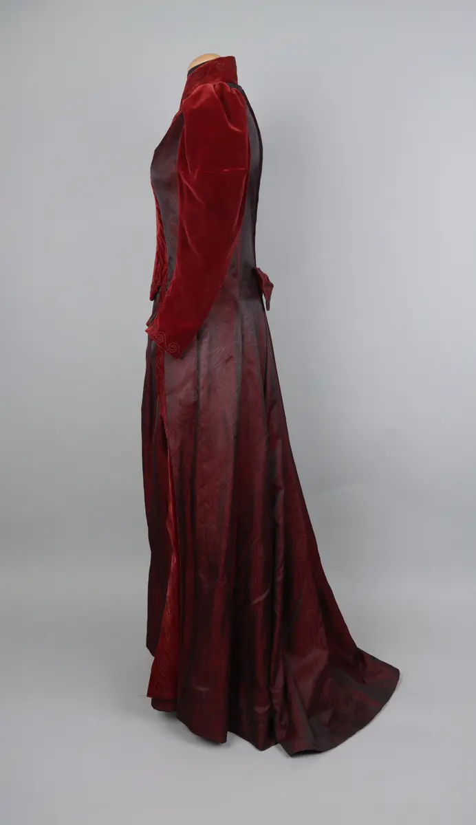 Klänning, av modell från 1891. Festklänning för dam av vinröd sammet och moire-liknande tyg. Sitter ihop i ett stycke med kjol och robe/överdel sammansatta. Stängs i ryggen med hyskor och hakar. Den höga kragen knäpps på med tryckknappar. Klänningens kjol har ett kortare släp.

