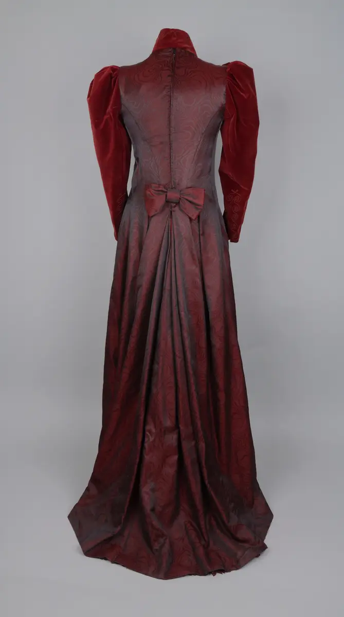 Klänning, av modell från 1891. Festklänning för dam av vinröd sammet och moire-liknande tyg. Sitter ihop i ett stycke med kjol och robe/överdel sammansatta. Stängs i ryggen med hyskor och hakar. Den höga kragen knäpps på med tryckknappar. Klänningens kjol har ett kortare släp.


