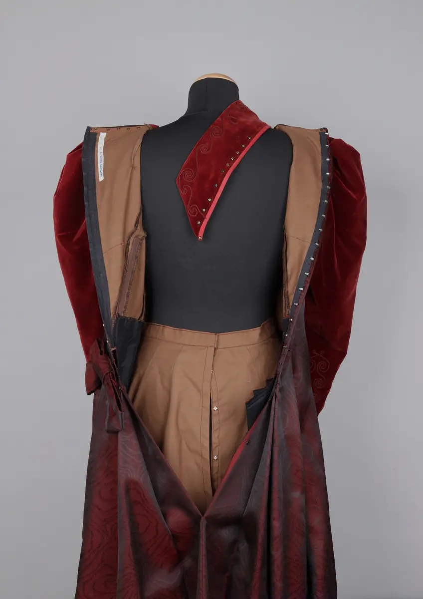 Klänning, av modell från 1891. Festklänning för dam av vinröd sammet och moire-liknande tyg. Sitter ihop i ett stycke med kjol och robe/överdel sammansatta. Stängs i ryggen med hyskor och hakar. Den höga kragen knäpps på med tryckknappar. Klänningens kjol har ett kortare släp.

