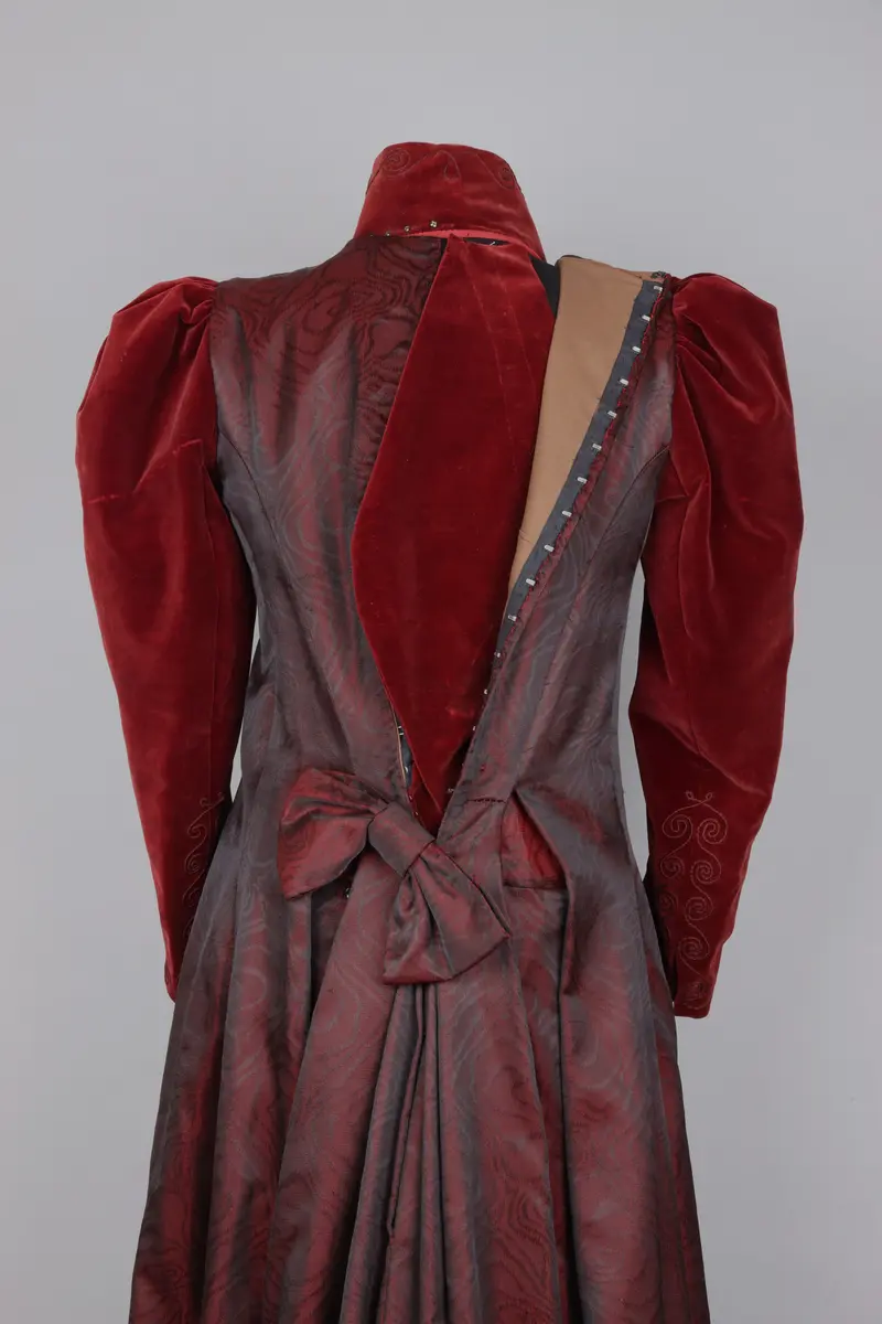 Klänning, av modell från 1891. Festklänning för dam av vinröd sammet och moire-liknande tyg. Sitter ihop i ett stycke med kjol och robe/överdel sammansatta. Stängs i ryggen med hyskor och hakar. Den höga kragen knäpps på med tryckknappar. Klänningens kjol har ett kortare släp.

