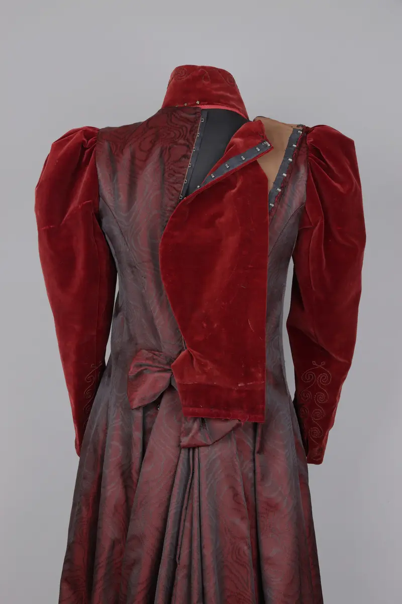 Klänning, av modell från 1891. Festklänning för dam av vinröd sammet och moire-liknande tyg. Sitter ihop i ett stycke med kjol och robe/överdel sammansatta. Stängs i ryggen med hyskor och hakar. Den höga kragen knäpps på med tryckknappar. Klänningens kjol har ett kortare släp.

